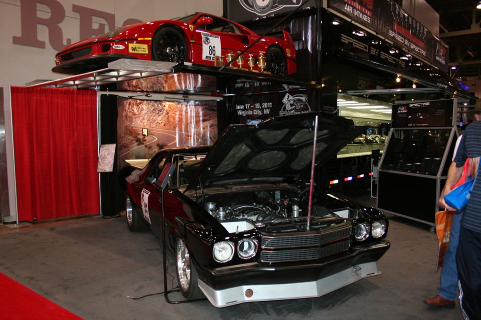 Black muscle car with raised hood beside a red sports car on an overhead display rack at an auto show