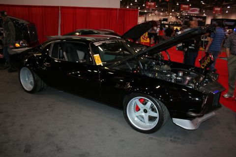 1998 Ford Crown Victoria - black custom fastback muscle car with wide white-faced wheels and an open hood is shown in profile on a show floor crowded with spectators.