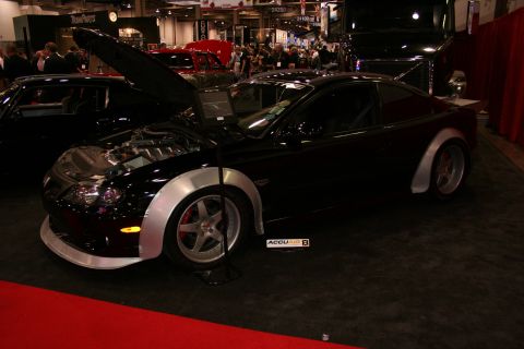 1998 Ford Crown Victoria - black coupe with raised hood, bolt-on silver fender flares and red brake calipers sits over a red carpet at a crowded indoor car show.