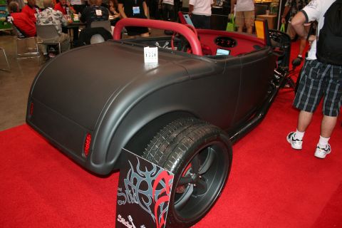 1998 Ford Crown Victoria - matte-black hot rod roadster with a red interior and a flame-decal display panel sits on red carpet among spectators at an indoor show.