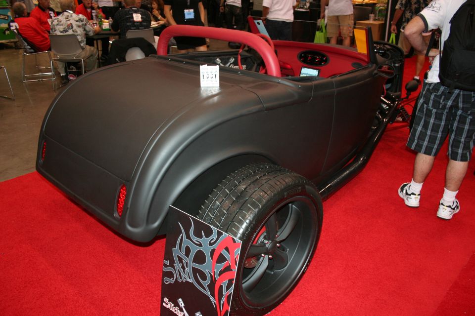 Matte-black hot rod roadster with red interior on a red carpet at an indoor auto show