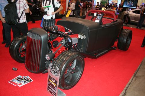 1998 Ford Crown Victoria - matte-black hot rod roadster reveals an exposed red-painted engine and chrome wheels, displayed on a red show floor with an info placard.