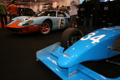 1998 Ford Crown Victoria - light-blue and orange Gulf-liveried race coupe numbered 6 sits behind a blue open-wheel race car numbered 34 at an indoor motorsport displa…