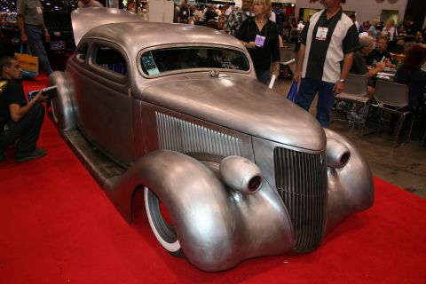 1998 Ford Crown Victoria - bare-metal chopped vintage coupe hot rod with a louvered hood and whitewall tires sits on red carpet surrounded by onlookers at a show.