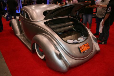 1998 Ford Crown Victoria - rear of a bare-metal chopped vintage coupe shows an open trunk and an orange license plate reading BLO FANG on a red show carpet.