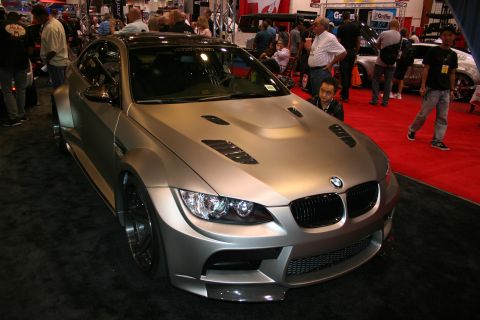 1998 Ford Crown Victoria - silver matte-finish BMW coupe with hood vents and a wide body kit sits on red carpet as spectators walk past at an indoor auto show.