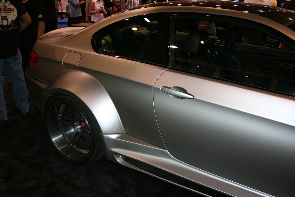 Side profile of a silver matte coupe with wide fenders and dark five-spoke wheels at an auto show