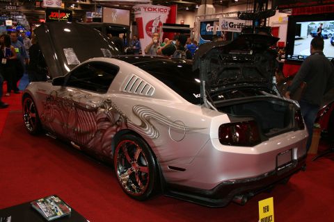 1998 Ford Crown Victoria - silver custom muscle coupe with swirling airbrushed graphics and an open rear hatch sits on red carpet beside a video screen at a show.