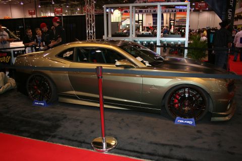 1998 Ford Crown Victoria - olive-green custom coupe with a wide body kit and dark wheels sits behind a rope barrier over red carpet at a busy indoor show.