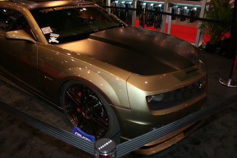 1998 Ford Crown Victoria - front three-quarter of an olive-gold custom coupe shows a black hood stripe and dark split-spoke wheels behind a barrier on a show floor.
