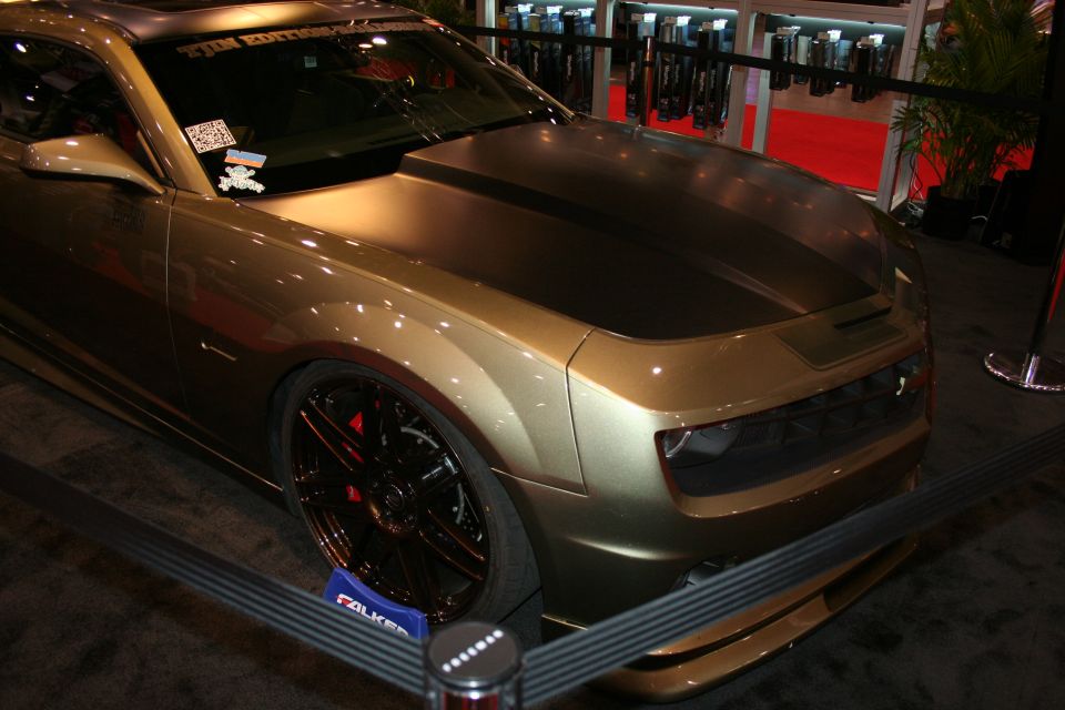 Front three-quarter of an olive-gold custom coupe with black hood and dark split-spoke wheels at a show