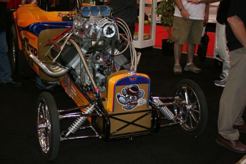 1998 Ford Crown Victoria - yellow vintage front-engine dragster with an exposed supercharged engine and chrome zoomie headers sits among spectators at an indoor show.
