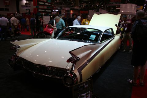1998 Ford Crown Victoria - cream-colored late-1950s Cadillac with chrome bullet bumpers and tall tailfins sits with its hood open as crowds gather at an indoor show.