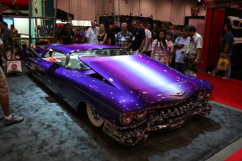 1998 Ford Crown Victoria - purple metalflake custom Cadillac with a chopped roof, whitewall tires and elaborate chrome sits on a show floor surrounded by spectators.