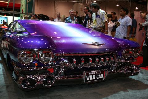 1998 Ford Crown Victoria - Front three-quarter of a purple metalflake custom Cadillac with heavy chrome and a license plate reading WILD CAD on a crowded show floor.