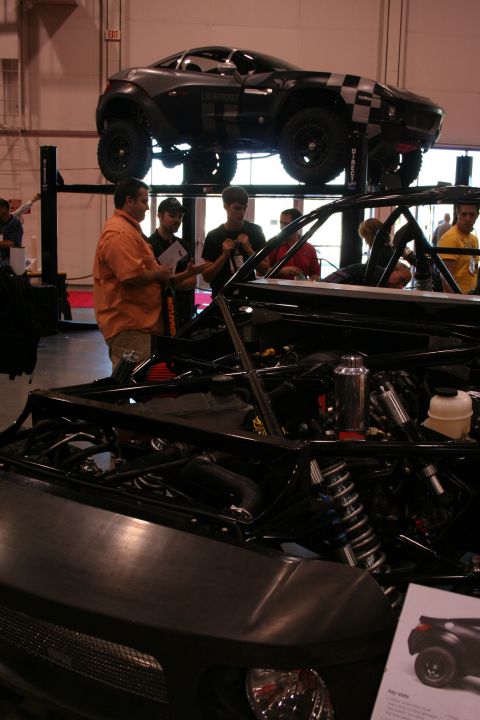 1998 Ford Crown Victoria - tube-frame off-road vehicle chassis with exposed coilovers sits at floor level while a camo-wrapped vehicle is raised on a lift behind it.
