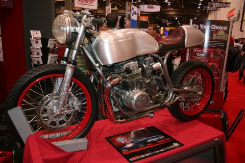 1998 Ford Crown Victoria - custom cafe-racer motorcycle with a polished bare-metal tank and tail, red-rimmed wheels and an exposed inline engine on a red show stand.