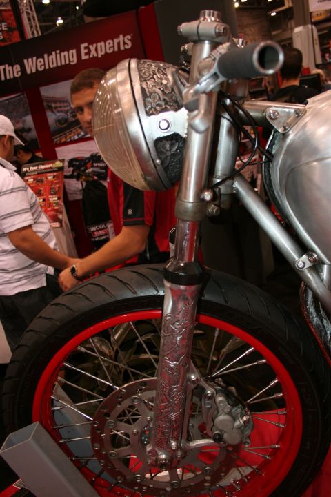 1998 Ford Crown Victoria - Front end of a custom motorcycle showing engraved fork legs, a round headlight and a red-rimmed front wheel at an indoor booth.