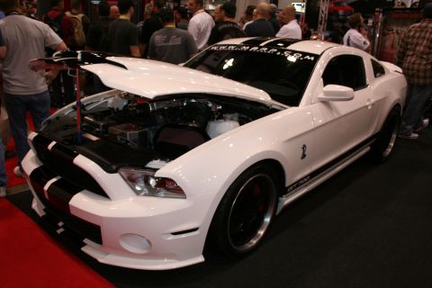 1998 Ford Crown Victoria - white Ford Shelby GT500 Mustang with black racing stripes and open hood revealing the engine, displayed before a crowd at an indoor show.