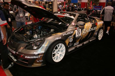 1998 Ford Crown Victoria - heavily liveried Lexus race car numbered 26 with open hood showing the engine and white wheels, on display at an indoor motorsports show.