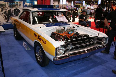 1998 Ford Crown Victoria - yellow-and-white Mr. Norm's vintage Dodge drag car with an exposed chrome V8 and dual carburetors, displayed on a blue show stand.