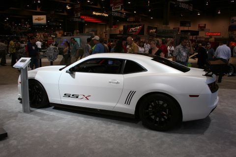 1998 Ford Crown Victoria - white Camaro coupe with 'SSX' side graphics and black wheels displayed before a crowd on a trade-show floor.