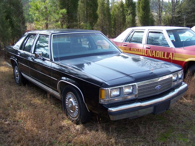 1991 Ford Crown Victoria parked next to a red fire rescue vehicle. The Crown Victoria is black with chrome trim