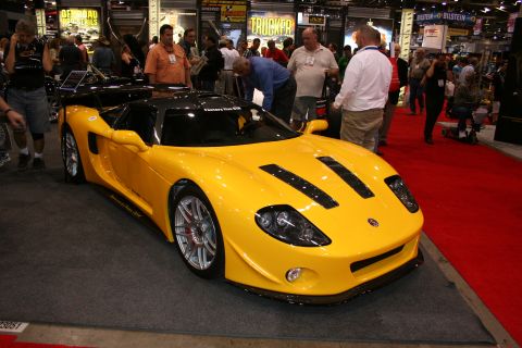 1998 Ford Crown Victoria - glossy yellow mid-engine sports car with hood vents and silver wheels displayed among attendees on a red show-floor carpet.