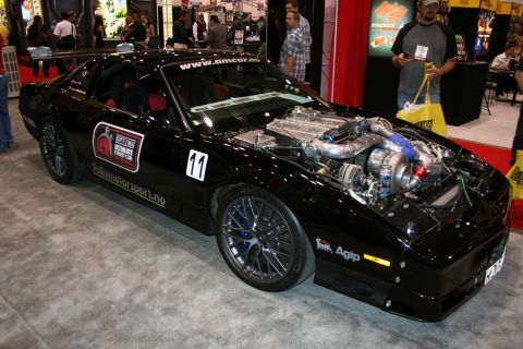 1998 Ford Crown Victoria - black number-11 race coupe with sponsor decals and an exposed turbocharged engine on display under bright trade-show lights.