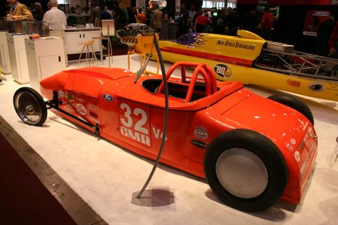 1998 Ford Crown Victoria - orange streamlined hot-rod lakester numbered 32 with an exposed V8 engine displayed beside a yellow streamliner at a show.