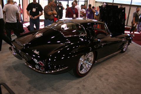 1998 Ford Crown Victoria - glossy black mid-1960s Corvette Sting Ray coupe with chrome wheels sits rear-quarter to the camera on red carpet amid show crowds.