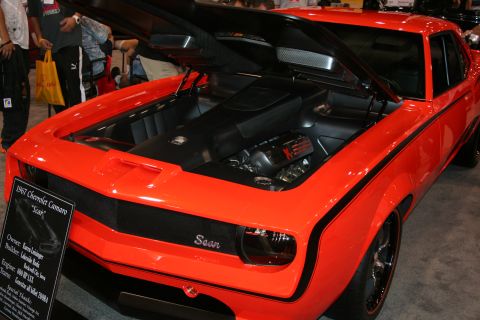 1998 Ford Crown Victoria - orange first-generation Camaro named Scar sits with hood open showing its engine, beside an info placard at an indoor show.