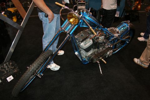 1998 Ford Crown Victoria - blue custom hardtail chopper with long springer front end and a brass headlight stands on black carpet at an indoor show.