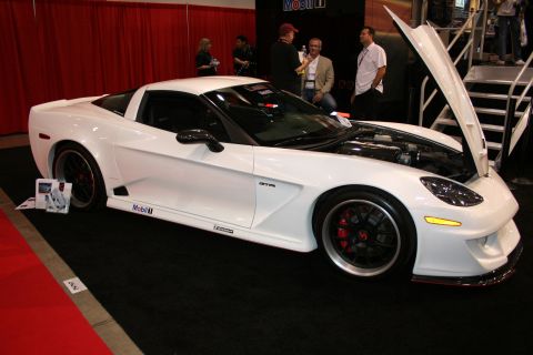 1998 Ford Crown Victoria - white widebody Corvette coupe with raised hood and red brake calipers, shown in profile on a carpeted booth floor.