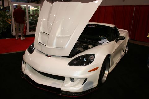1998 Ford Crown Victoria - white widebody Corvette coupe with raised hood and vented bonnet, front three-quarter on black carpet at an indoor show.