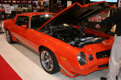 1998 Ford Crown Victoria - orange second-generation Camaro coupe with hood and hatch open, displayed on red carpet against a vendor backdrop.