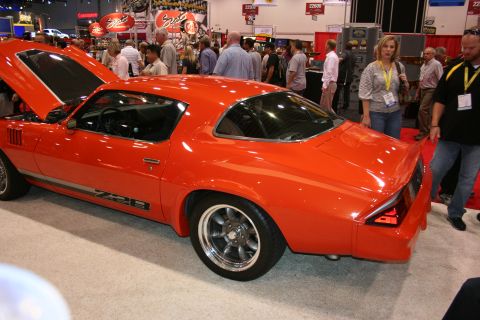 1998 Ford Crown Victoria - orange second-generation Camaro Z28 coupe on chrome wheels, shown rear-quarter amid show attendees on the floor.