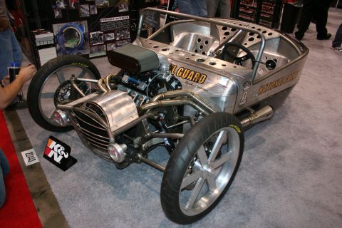 1998 Ford Crown Victoria - bare-aluminum open-wheel custom roadster with an exposed engine, swept exhaust and National Guard lettering sits on red carpet at a show.