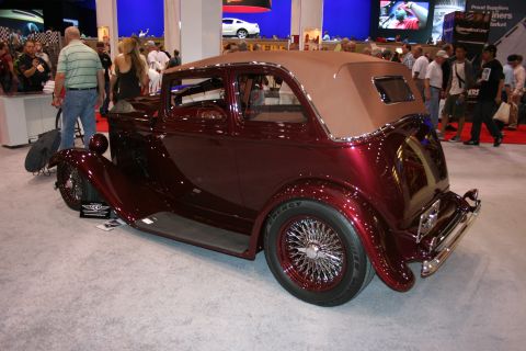 1998 Ford Crown Victoria - deep-maroon vintage sedan hot rod with a tan folding top and chrome wire wheels sits on display at an indoor show.