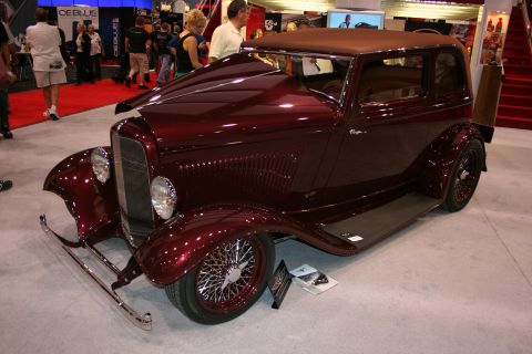 1998 Ford Crown Victoria - glossy maroon 1930s-style coupe hot rod with chrome wire wheels and side-exit exhaust sits on a show stand.