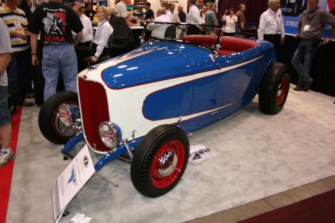 1998 Ford Crown Victoria - blue-and-white highboy roadster hot rod with red wire wheels and a red interior sits on display at an indoor show.