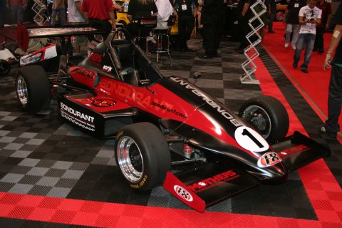 1998 Ford Crown Victoria - black-and-red open-wheel formula-style race car numbered 1 with Bondurant lettering sits on a red-and-black checkered show floor.