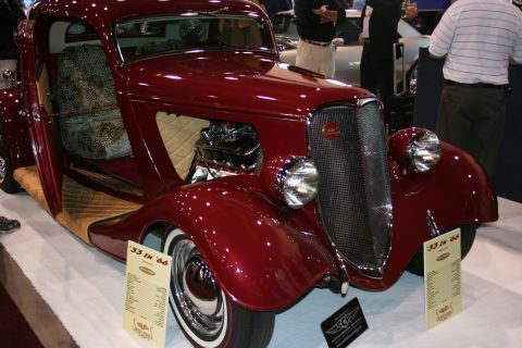 1998 Ford Crown Victoria - deep-maroon vintage coupe hot rod with chrome headlights and a polished engine sits beside spec cards reading '33 in 66' at a show.