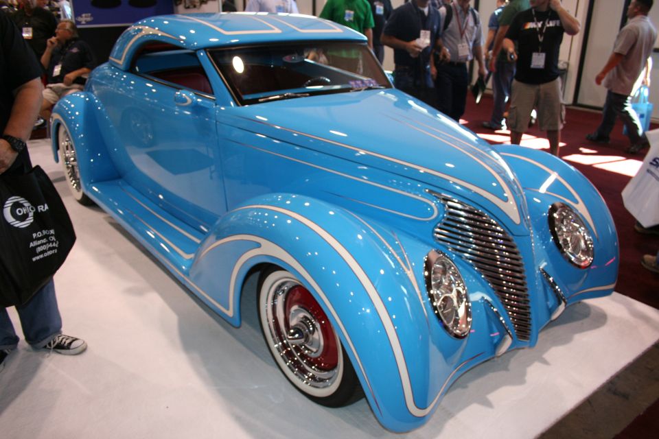 Front three-quarter of a sky-blue custom coupe with pinstriping, chrome wheels and red rims at an indoor show