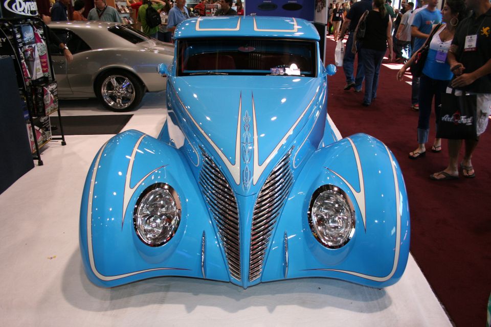 Head-on front view of a sky-blue custom coupe with V-shaped chrome grille and pinstriping at a show
