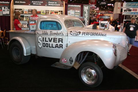 1998 Ford Crown Victoria - silver vintage gasser-style pickup lettered 'Silver Dollar' sits with a raised front axle and zoomie headers at an indoor show.