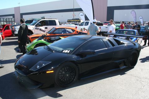 1998 Ford Crown Victoria - black Lamborghini supercar with a raised rear wing and black wheels parked among other vehicles at an outdoor show.