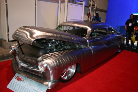 1998 Ford Crown Victoria - bare polished-metal chopped custom lead-sled coupe sits low on a red carpet with its hood open inside a show hall.