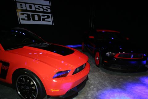 1998 Ford Crown Victoria - red and a black Ford Mustang coupe sit side by side on a dimly lit stage beneath an illuminated Boss 302 sign.