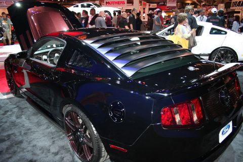 1998 Ford Crown Victoria - Rear three-quarter of a dark blue Ford Mustang coupe with rear-window louvers and dark wheels in a busy indoor show aisle.